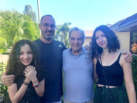 Four family members smiling warmly together in a lush backyard under blue skies.