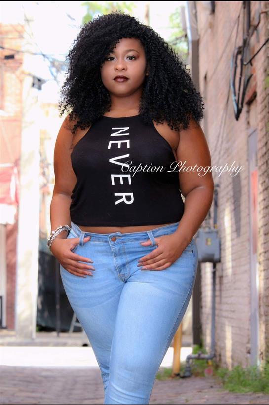 A woman in a black tank top and blue jeans strolls confidently in an urban alleyway.