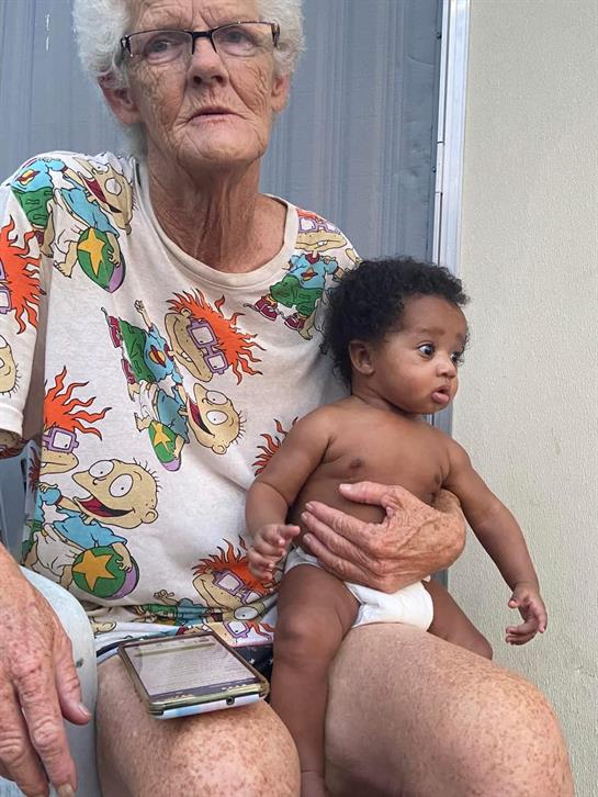 An elderly woman in casual clothing sits outside holding a baby who looks curious and attentive.