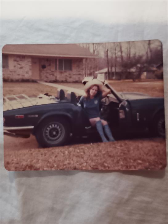 A woman sits on the edge of a convertible car in front of a house surrounded by fallen leaves.