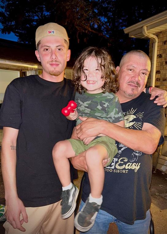 A man holds a smiling child while another man stands beside them in an outdoor setting.