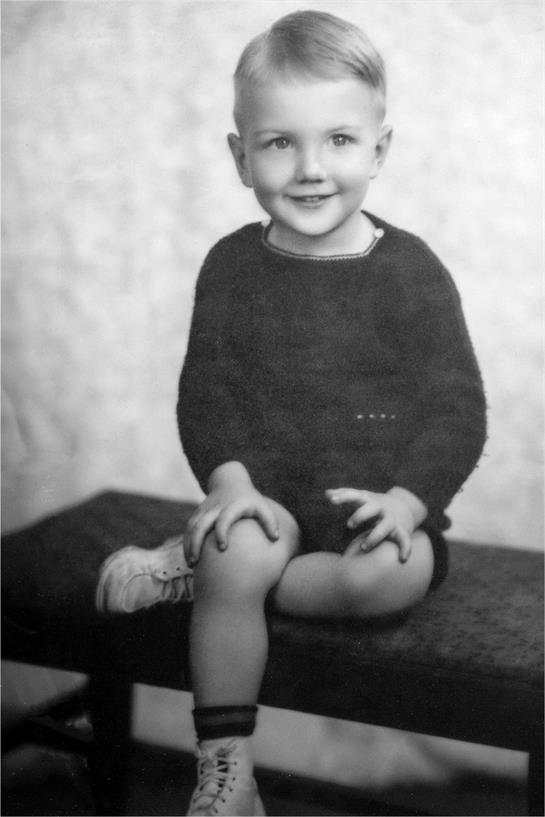 A cheerful young boy with light hair poses on a bench wearing a dark outfit and sneakers.