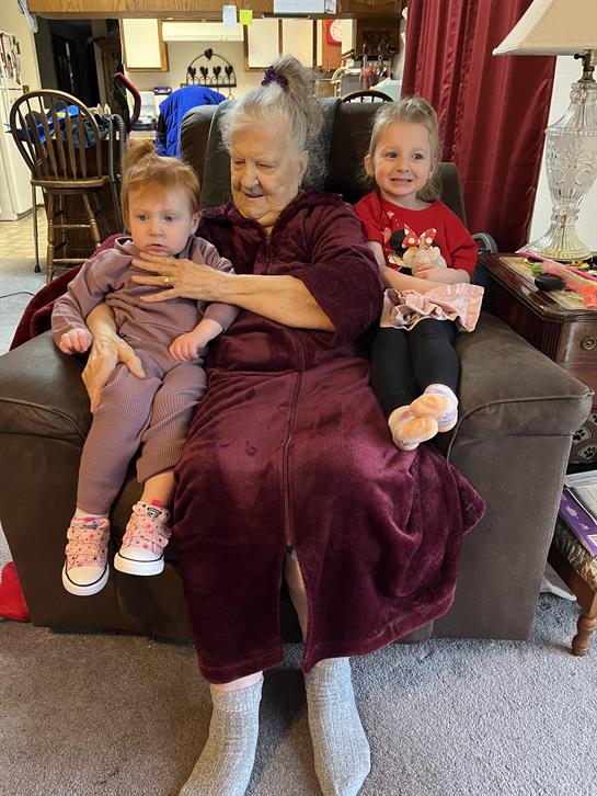 Elderly woman sits on a couch with two small girls, sharing smiles and a warm moment together.