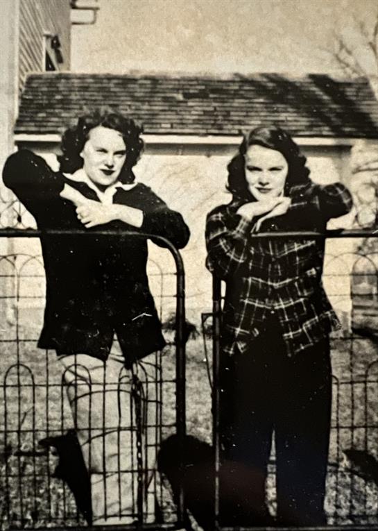 Two women lean against a fence in a rural setting, showcasing vintage fashion styles and smiles.
