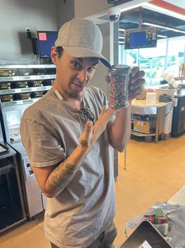 Individual shows a jar containing assorted colorful items while smiling in a cafe.