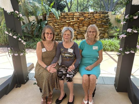 Three women sit together, celebrating a memorable occasion in a serene garden with vibrant plants.