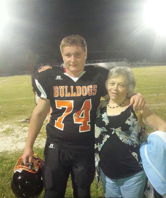 A young football player in uniform poses with his grandmother after the game under bright lights.