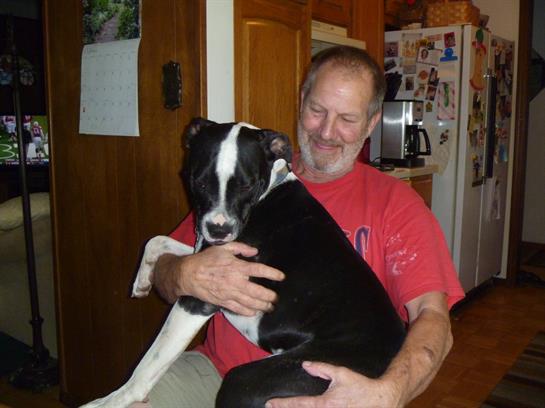 A man embraces his large black and white dog in a warm kitchen atmosphere, smiling happily.