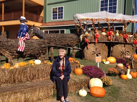 An elderly woman sits beside colorful pumpkins and hay bales during a vibrant autumn festival.
