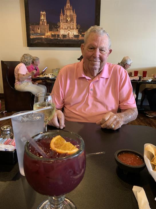 An elderly man smiles while sitting at a table with a large drink and salsa in front of him.