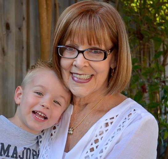 A cheerful grandmother embraces her young grandson, both smiling happily amid green foliage.