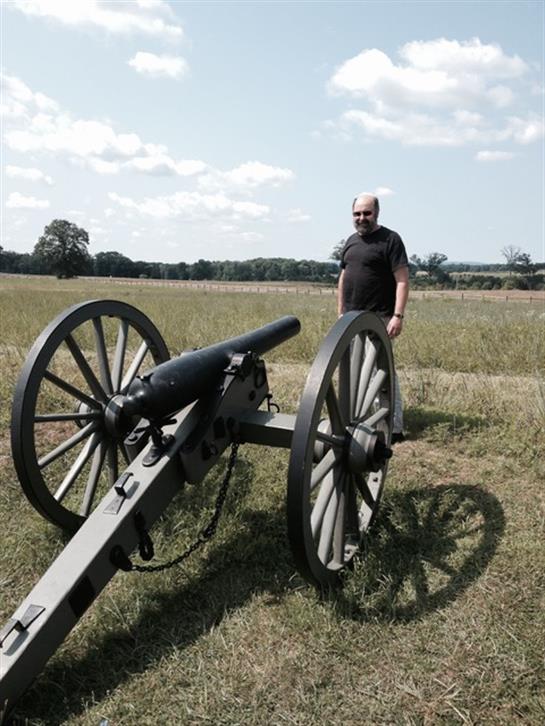 A man stands next to a large, historic cannon in an open field under a blue sky.