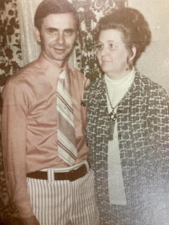 A couple stands together smiling in vintage attire, surrounded by decorative curtains.