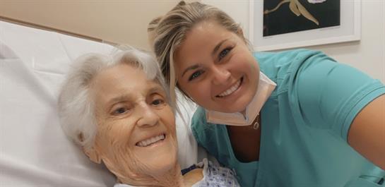 A nurse poses with an elderly patient, both smiling in a bright hospital room, showcasing care.