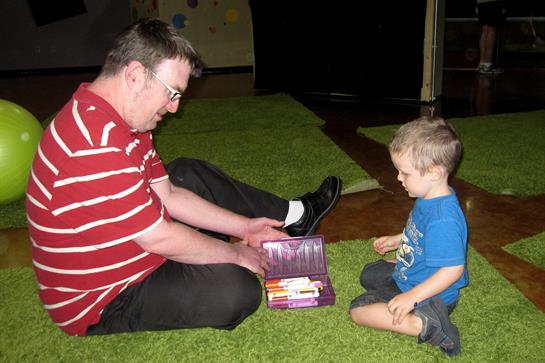 A man and a young boy are seated on a green carpet, exploring toys from a box together.