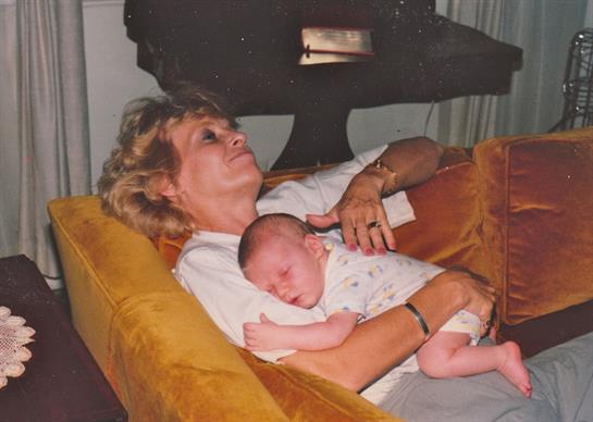 An elderly woman relaxes on a sofa while holding a sleeping baby, exuding warmth and love.