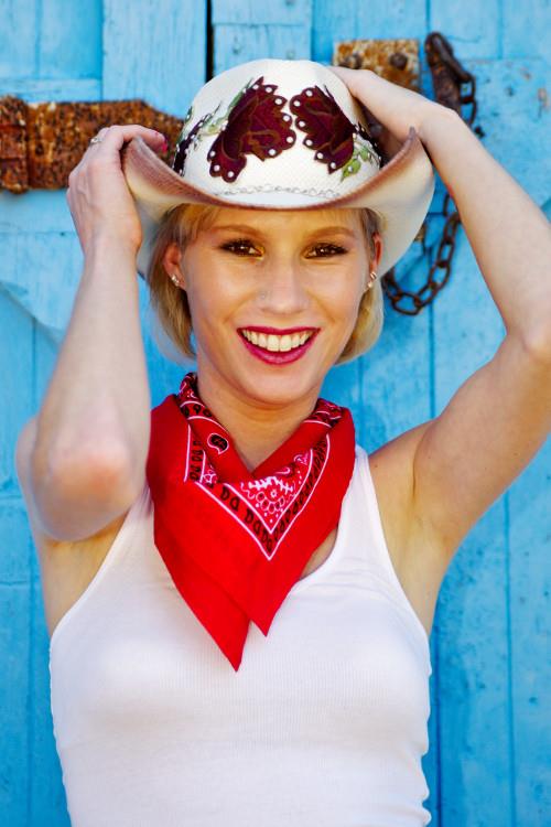 Woman with blonde hair adjusts her cowboy hat while smiling against a blue rustic backdrop.