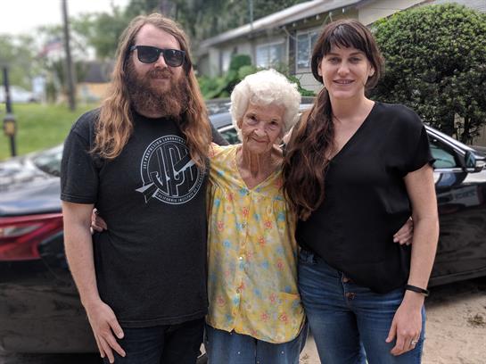 Three individuals stand together outside a house, sharing smiles and laughter in the warm sun.