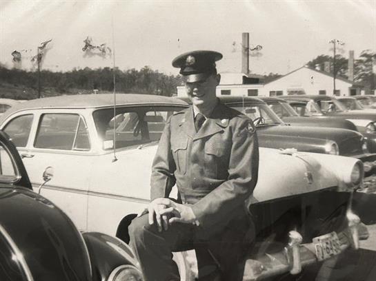 A soldier in uniform kneels next to classic cars at a military base, showcasing post-war life.