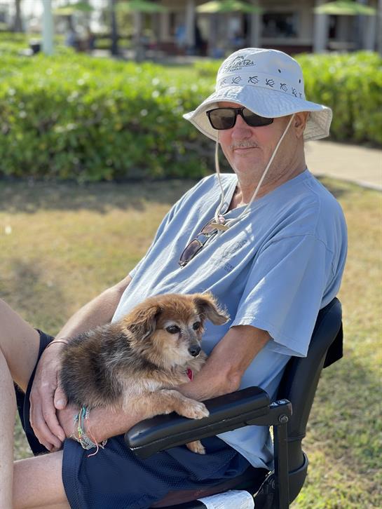 A man wearing sunglasses and a hat relaxes outdoors with a small dog on his lap.