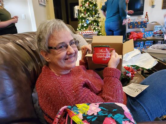 A woman happily unwraps a present while sitting on a couch surrounded by family.