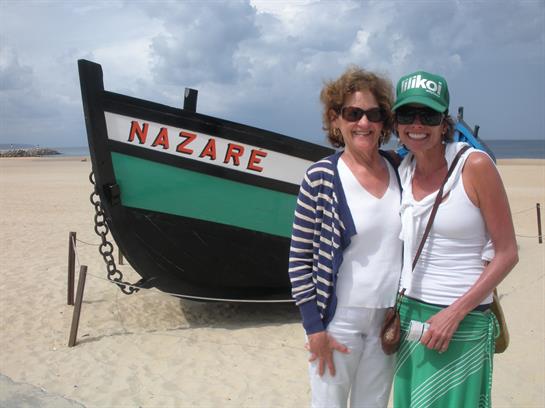 Two women smile and pose in front of a colorful fishing boat on a sandy beach.