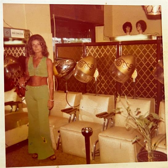 A woman stands in a vintage salon, showcasing retro decor and hair styling tools.