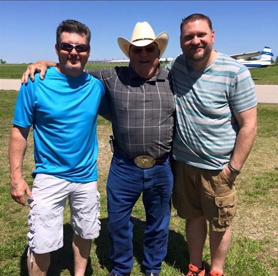 Two friends stand with a cowboy in a sunny field near an airstrip, enjoying a cheerful moment.