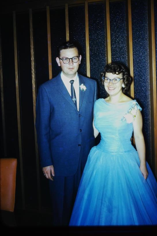 A smiling couple stands close together, dressed elegantly for a festive occasion.