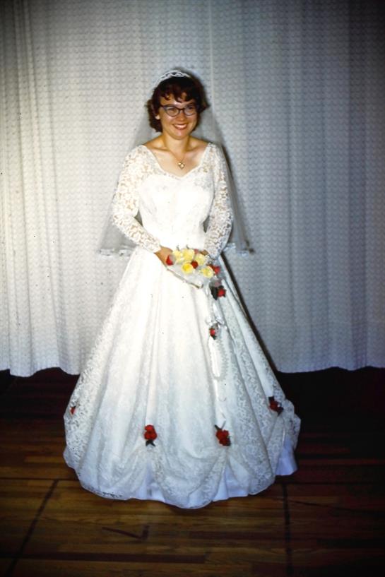 A bride stands proudly in a stunning white dress adorned with lace and roses.