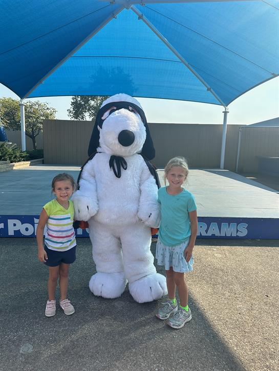 Two children pose with a large cartoon character under a blue shade in a park setting.
