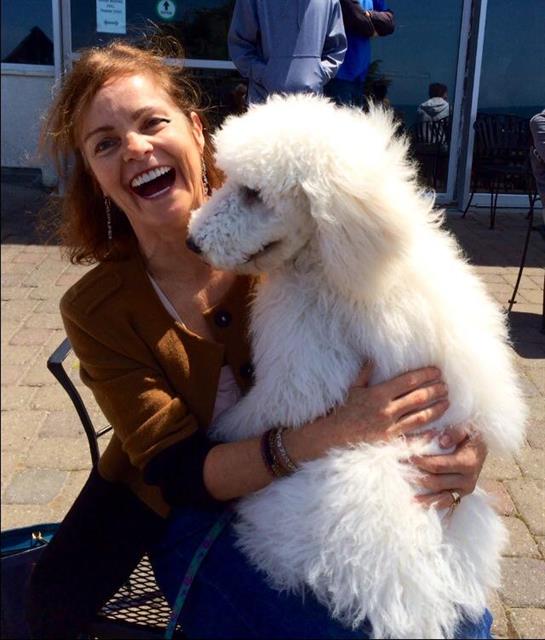 A woman laughs while holding her fluffy white dog in a vibrant outdoor area during daytime.