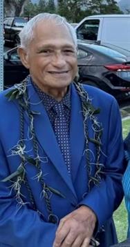 An elderly man in a blue suit and lei smiles warmly while attending a festive gathering outside.