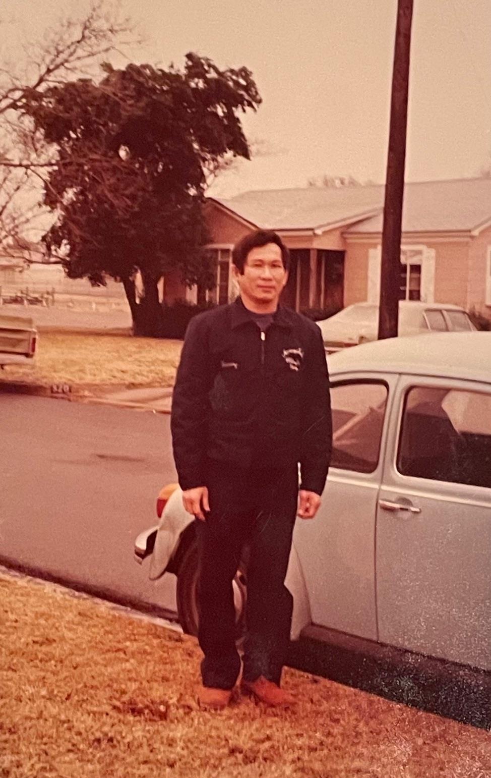 A man wearing a dark outfit stands next to a vintage car in a quiet neighborhood.