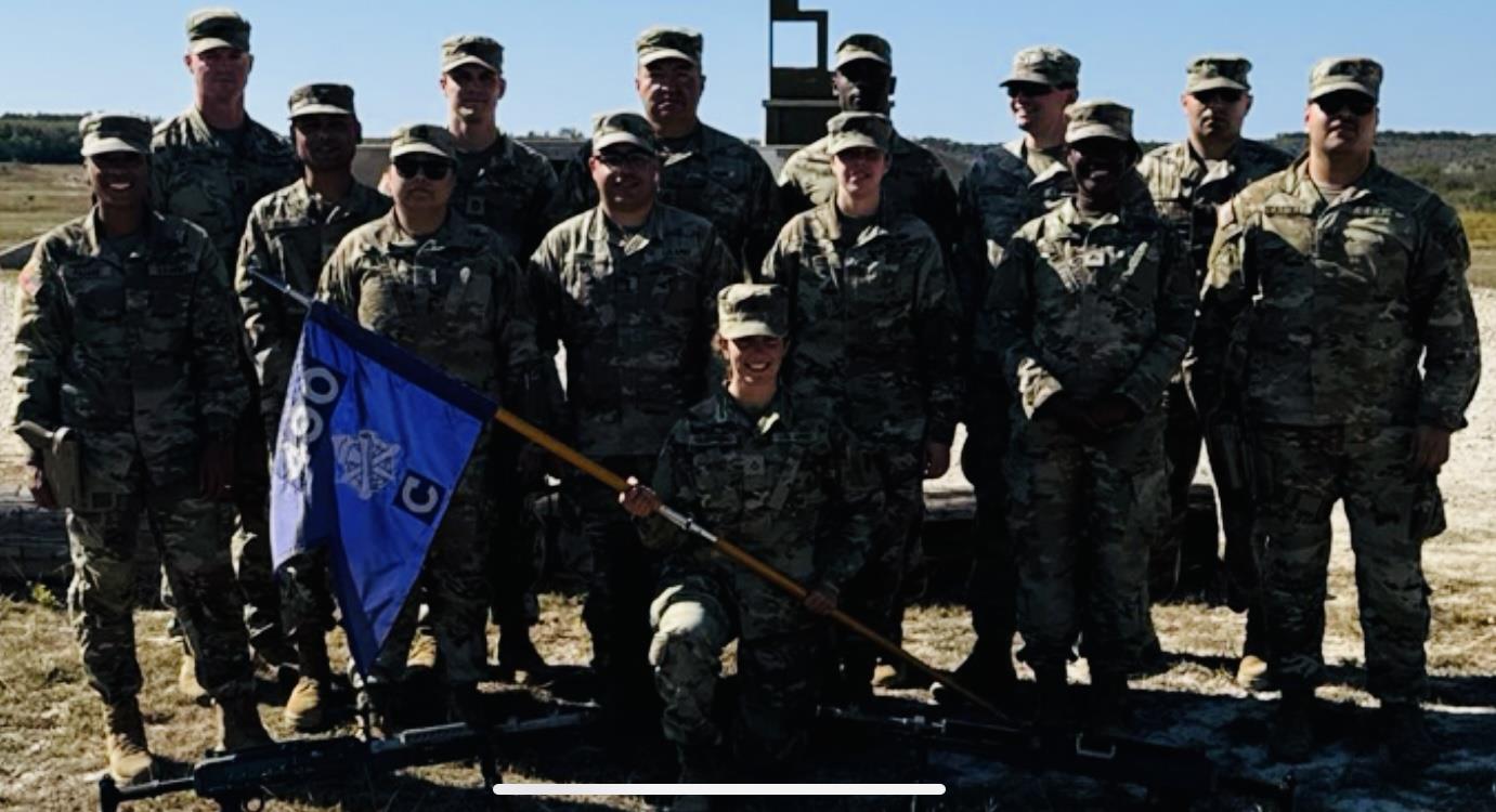 Soldiers dressed in camouflage uniforms gather for a group photo outdoors with a flag.