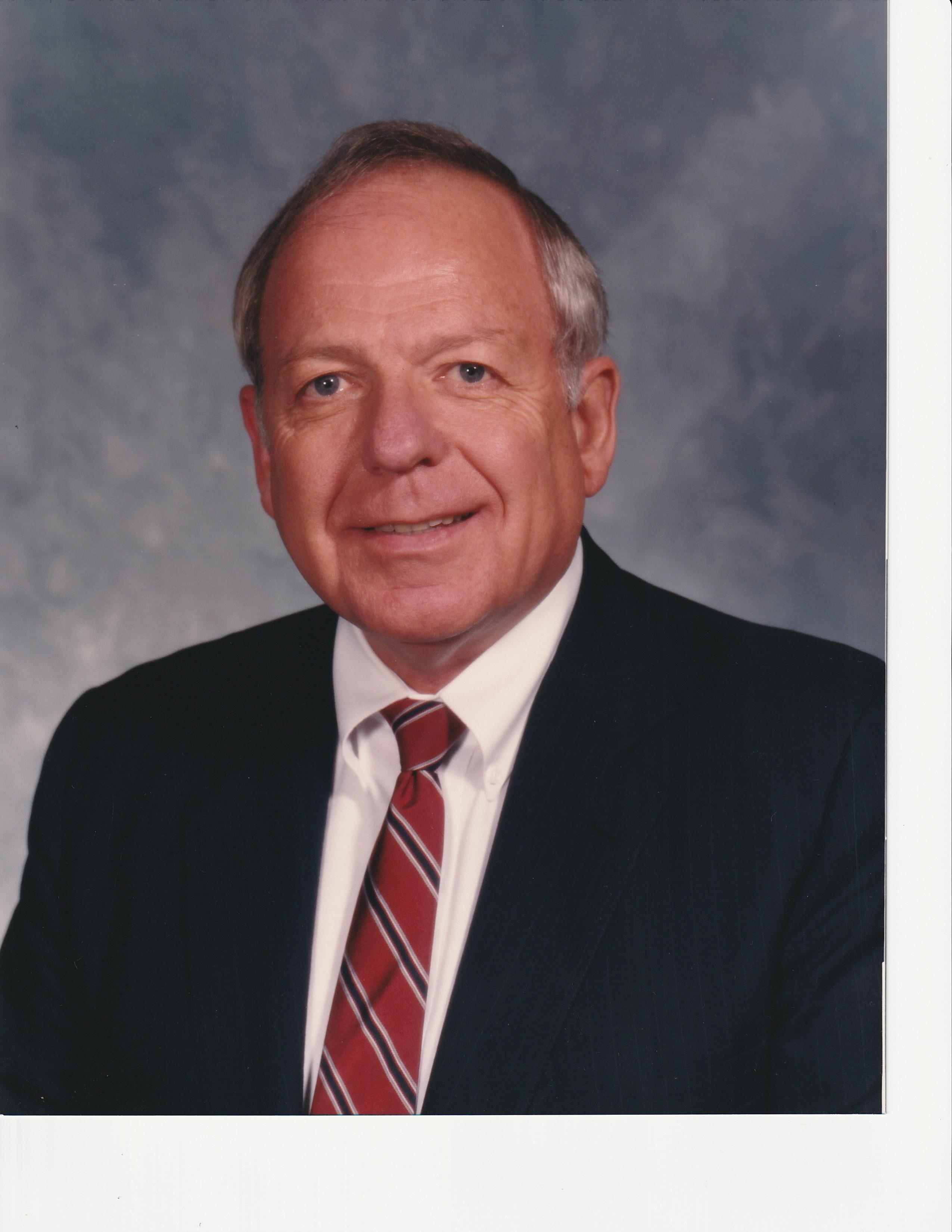 A well-dressed older man smiles warmly while posing against a neutral backdrop.
