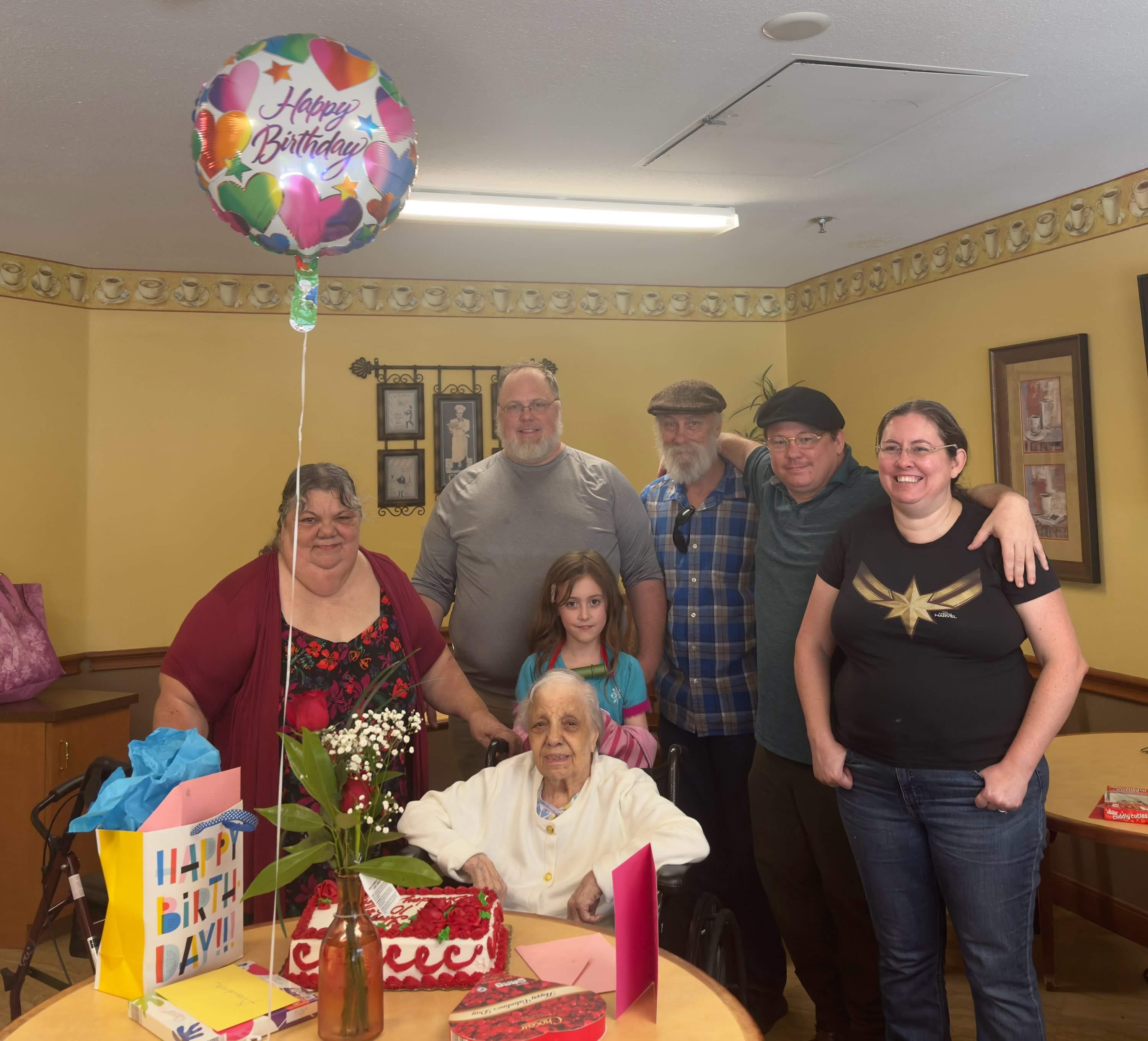 Family celebrates an elderly man's birthday with balloons and gifts in a cozy setting.