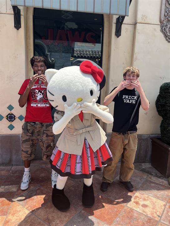 Two young men stand beside a large character dressed in a fashionable outfit, smiling joyfully.