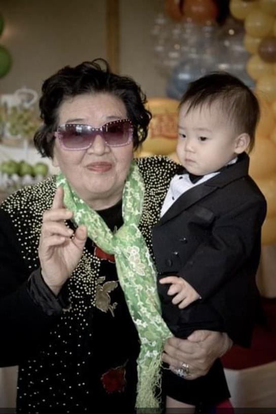 Elderly woman in sunglasses proudly holds a formally dressed child at a festive gathering.