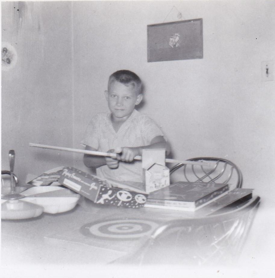 A boy sits at a table, holding a toy sword, surrounded by games and toys in the kitchen.