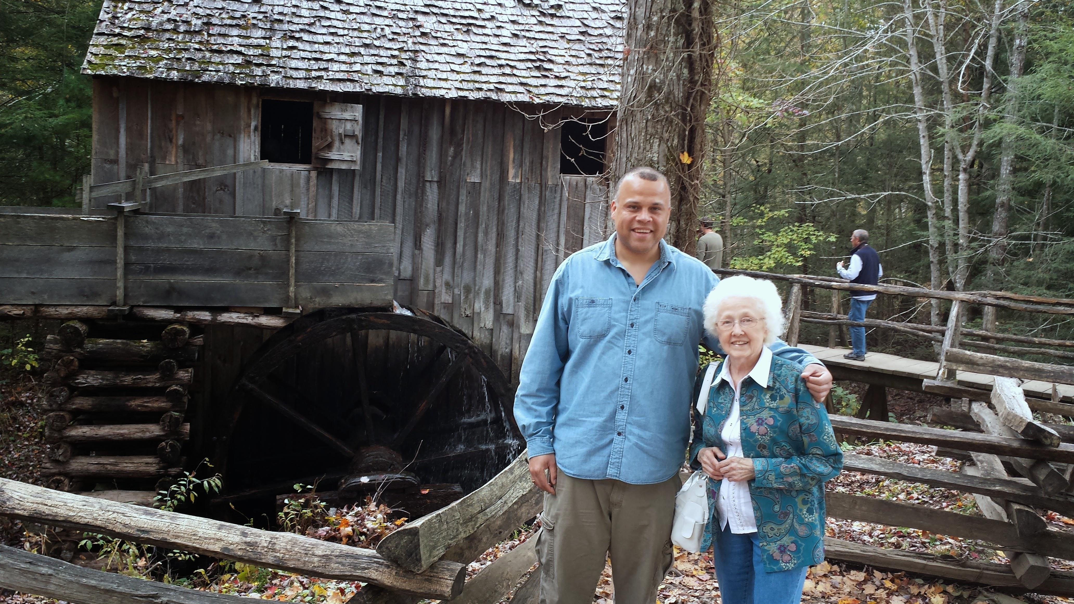 Two individuals stand smiling in front of a rustic watermill surrounded by colorful autumn foliage.