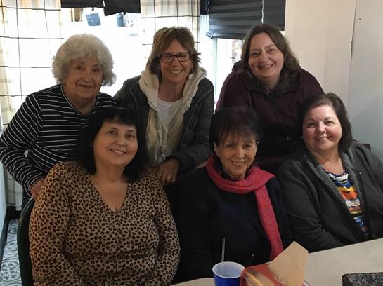 Six women gather around a table, sharing laughter and enjoying each other’s company indoors.