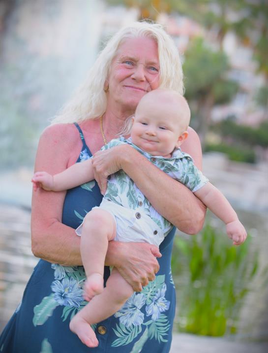 A joyful woman smiles while cradling a happy baby near a fountain in an outdoor park.