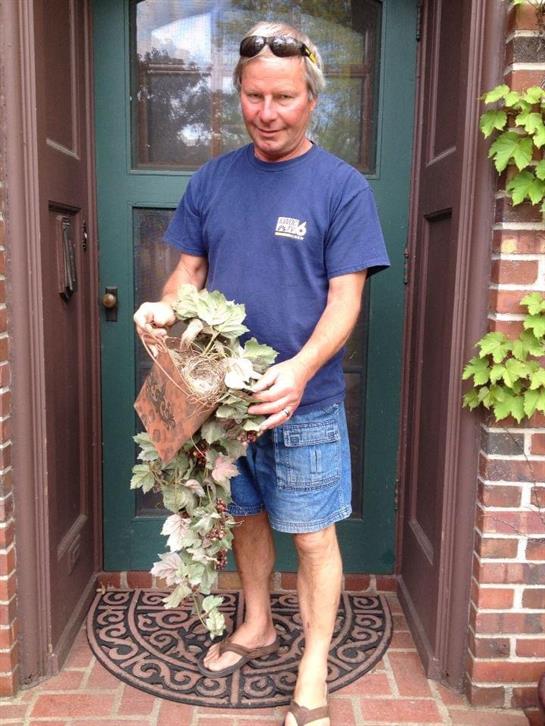 A man stands in front of a brick house, holding a decorative plant in his hands.