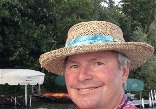 A cheerful man with a straw hat enjoys a sunny day by the lake, surrounded by boats.