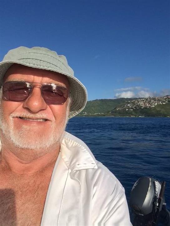 Elderly man with a beard smiles while resting on a boat in clear blue water under a sunny sky.