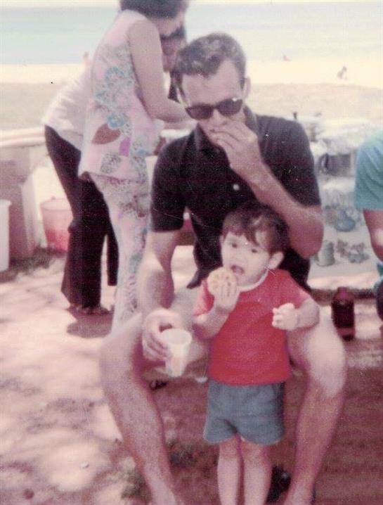 A man in sunglasses enjoys snacks with a child while others mingle nearby on a sunny day.