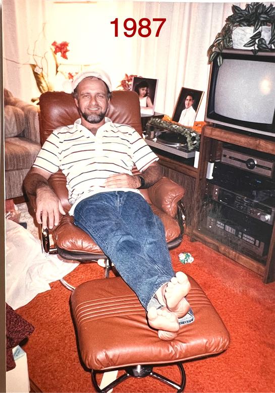 A man in a striped shirt reclines in a leather chair, surrounded by vintage decor in 1987.