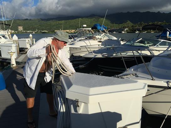A man adjusts ropes on a boat at a marina during daylight in a calm harbor.