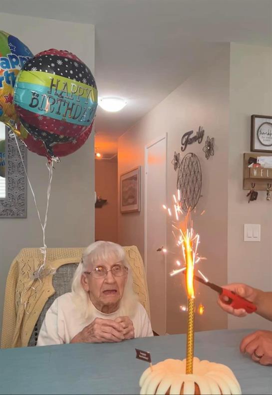 An elderly woman watches with joy as a sparkler is lit on her birthday cake, surrounded by balloons.